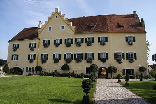 Das imposante Schloss mit seiner hellgelben Farbe und grünen Fensterläden. Davor geht ein gepflasterter Weg auf den Eingang zu. Rechts und links eingerahmt von grünem Gras.
