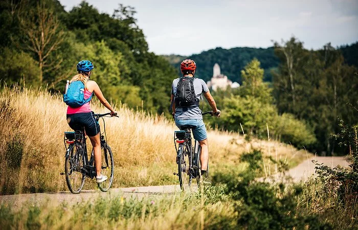 Zwei Radler, eine Frau und ein Mann, sind auf dem Radweg unterwegs. Die Gräser entlang des Radweges sind recht schon recht vertrocknet und braun. Ihr Ziel ist die Burg Kipfenberg die inmitten des Waldes in weiß bereits unscharf zu sehen ist.