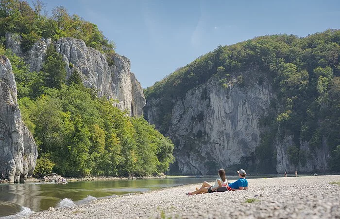 Ein Pärchen liegt am Kiesstrand und lässt seinen Blick zum Donaudurchbruch schweifen. Die Donau fließt zwischen rund 80 m hohen Felswänden hindurch.