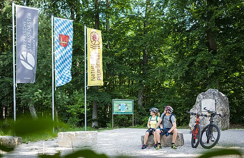 Geographischer Mittelpunkt Bayerns Zwei Radler sitzen auf einem Felsbrocken am Geographischem Mittelpunkt. Rechts von ihnen sind ihre zwei Fahrräder abgestellt. Hinter ihnen ist rechts eine Infotafel und links von ihnen sind drei Fahnen gehisst.