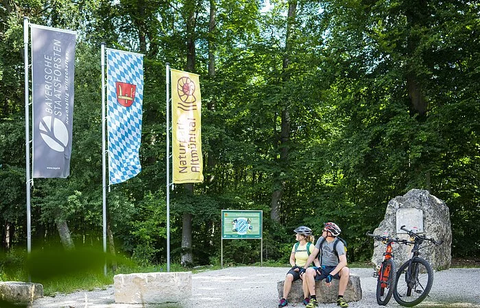 Zwei Radler sitzen auf einem Felsbrocken am Geographischem Mittelpunkt. Rechts von ihnen sind ihre zwei Fahrräder abgestellt. Hinter ihnen ist rechts eine Infotafel und links von ihnen sind drei Fahnen gehisst.