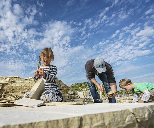 Ein Mann mit zwei Kindern suchen im Steinbruch nach Fossilien. Das Mädchen löst vorsichtig Gesteinsplatten mit Hammer und Meißel. Der Junge mit dem Mann schauen sich ihre Funde an.