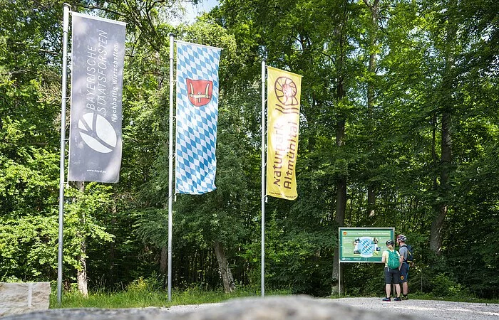 Zwei Wanderer stehen an der Infotafel am Geographischen Mittelpunkt Bayerns bei Kipfenberg und lesen interessiert. Links von ihnen sind drei Fahnen gehisst.