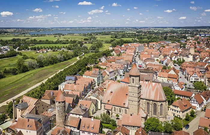 Luftaufnahme von Gunzenhausen am Altmühlsee. Im Vordergrund sind viele Häuser und der Kirchturm zu sehen, im Hintergrund grüne Wiesen, sowie strahlend blauer Himmel.