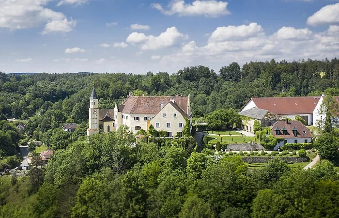 Das Schloss Hexenagger von oben zu sehen. Eingebettet in grünem Blätterkleid. Der Himmel ist hellblau und weiße Wolken sind zu sehen.