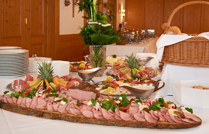 Es leckeres kaltes Buffet ist vor einem Fenster aufgebaut. Es gibt Platten mit Wurst, Lachs und Käse. Erhöht steht der große gut gefüllte Brotkorb. Eine Blumendeko und Teller gehören ebenfalls dazu.