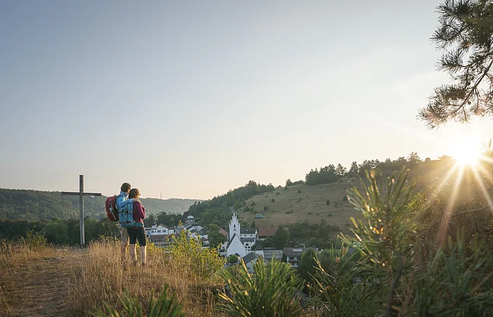 Zwei Wandernde stehen neben dem Kreuz auf dem Pfünzer Kirchberg und schauen über Pfünz zum Osterberg mit seiner Wacholderheide. Über dem Osterberg geht gerade die Sonne unter.