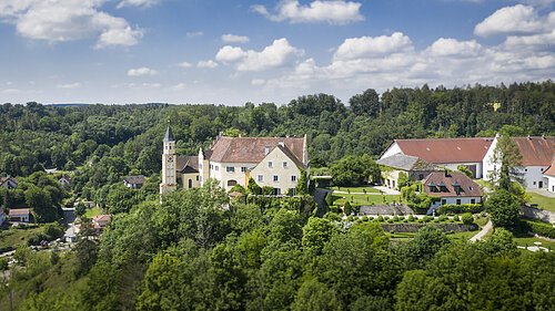 Das Schloss Hexenagger von oben zu sehen. Eingebettet in grünem Blätterkleid. Der Himmel ist hellblau und weiße Wolken sind zu sehen.