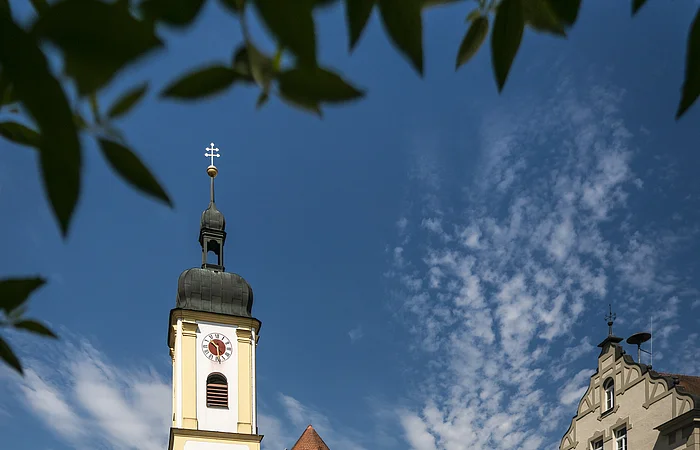 Kirche mit Turm und Uhr, umgeben von Gebäuden und Bäumen, unter blauem Himmel mit Wolken.