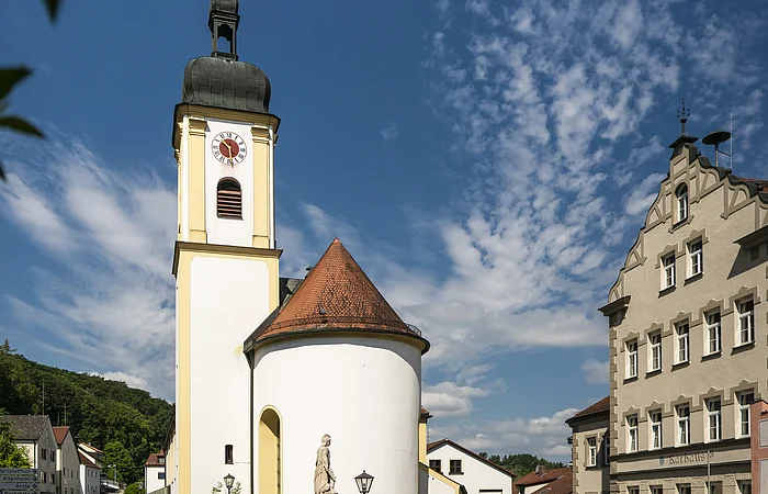 Heilig-Kreuz-Kirche in Altmannstein Kirche mit Turm und Uhr, umgeben von Gebäuden und Bäumen, unter blauem Himmel mit Wolken.