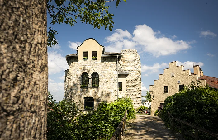 Steinernes Gebäude mit Rundturm und Weg mit Holzzaun bei sonnigem Himmel und Baum im Vordergrund