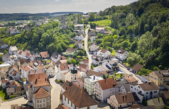 Luftaufnahme eines Dorfes mit Kirche, Wohnhäusern, bewaldeten Hügeln und einer Burgruine im Hintergrund.
