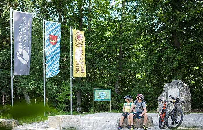 Zwei Radfahrer mit Helm sitzen auf Steinbank im Wald neben zwei Mountainbikes und drei Fahnen.