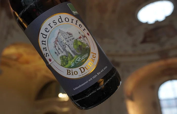 Flasche Sandersdorfer Bio Dunkel Bier mit Etikett, im Hintergrund unscharfe Architektur.